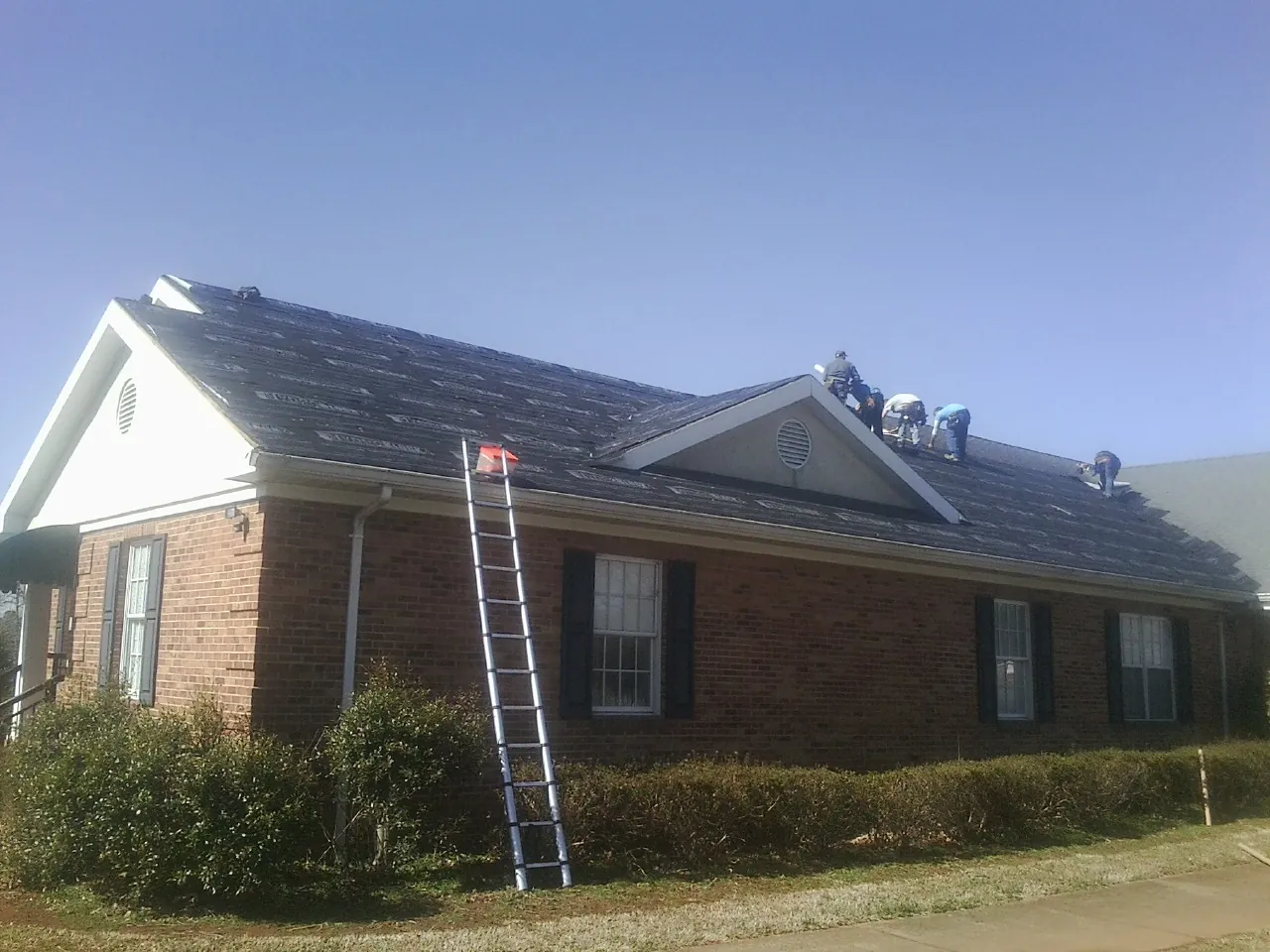 Crew tearing shingles off a brick ranch roof with a ladder set — Metal Roof Repair work by our crew in Snyder